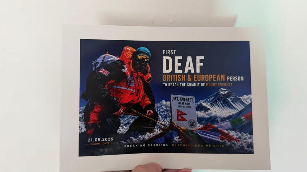 A climber in red gear holds a flag at Mt. Everest’s summit with the text: "First Deaf British & European person to reach the summit of Mount Everest. Summit date: 21.05.2026.