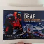 A climber in red gear holds a flag at Mt. Everest’s summit with the text: "First Deaf British & European person to reach the summit of Mount Everest. Summit date: 21.05.2026.