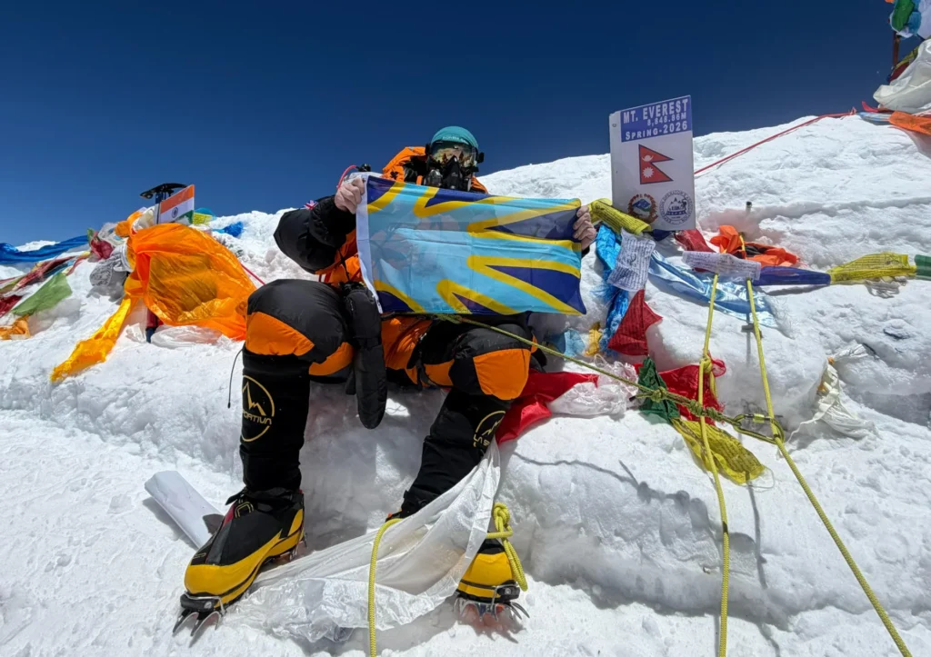 A climber in orange and black gear sits on snow at Mt. Everest’s summit, holding a blue and yellow flag, surrounded by colorful prayer flags and a summit sign.